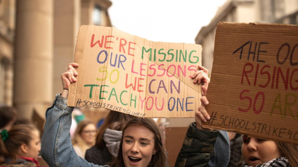 Mladé dievča drží banner s nápisom: ”Nie sme teraz na hodinách v škole, aby sme ťa mohli niečo naučiť.”