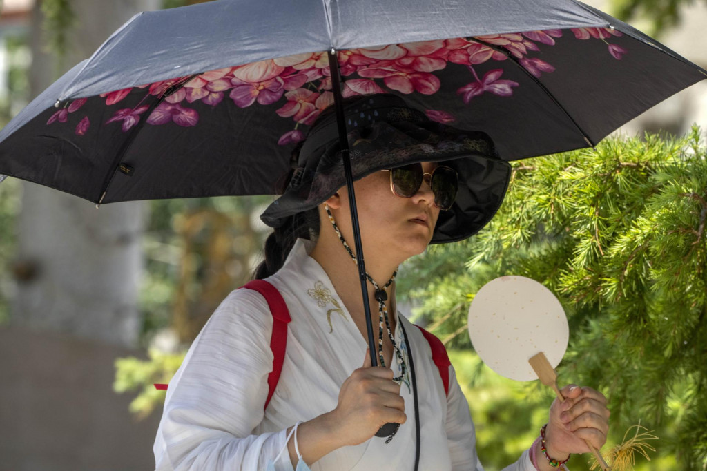 Žena kráča s dáždnikom a vejárom na osvieženie vo verejnom parku počas horúčav v Pekingu. FOTO: TASR/AP