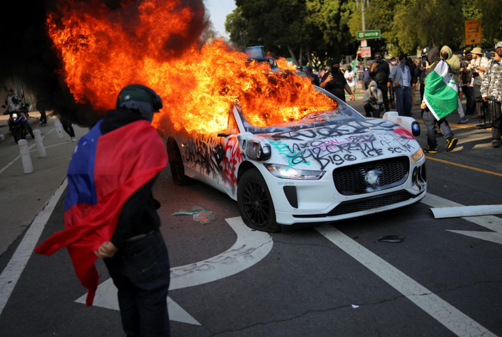 Demonštrant zahalený vo vlajke Samoy stojí pri horiacom aute počas protestu proti federálnym imigračným raziám v centre Los Angeles. FOTO: Reuters