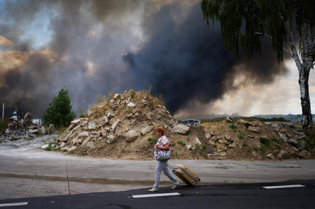 Žena kr&aacute;ča s kufrom po ceste okolo hust&eacute;ho čierneho dymu, ktor&yacute; st&uacute;pa z horiaceho skladu po ruskom dronovom &uacute;toku na Kyjev vo &scaron;tvrtok 27. augusta 2026. FOTO: TASR/AP