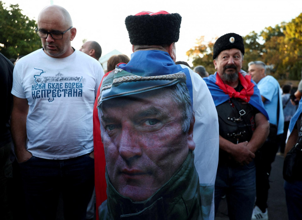 Muž s vlajkou vo farb&aacute;ch srbskej vlajky a s fotografiou b&yacute;val&eacute;ho bosniansko-srbsk&eacute;ho vojnov&eacute;ho gener&aacute;la Ratka Mladića stoj&iacute; medzi ľuďmi zhromažden&yacute;mi v Kostole sv. Luk&aacute;&scaron;a. FOTO: Reuters